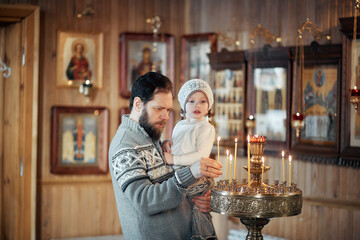 A Russian man with a beard and a daughter is standing in an Orthodox Church, lighting a candle and praying in front of the icon.