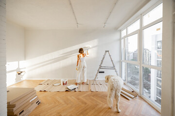 Woman with her dog paints the wall in white color, making repairment in newly purchased apartment....