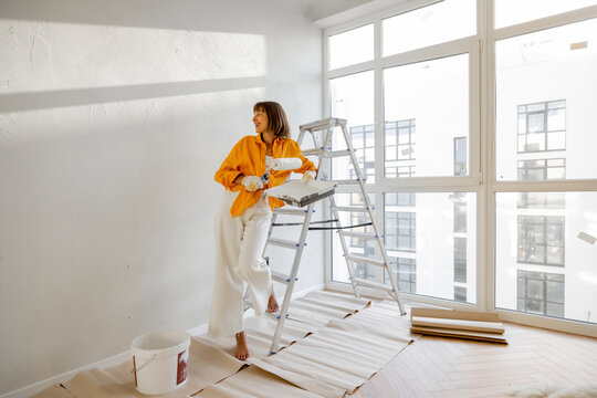 Portrait of a young woman making repairing in apartment, standing with paint roller near ladder in bright room of her new home