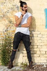 Young man with long dark hair and muscles hands posing in front of brick wall in urban part of city