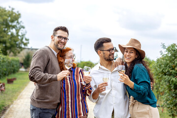 Group of four friends standing in vineyard with glasses of vine in hands, talk to each other and smile
