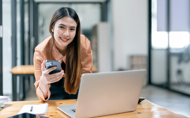 Charming Asian woman with a smile looking at camera standing holding mobile phone at the office.