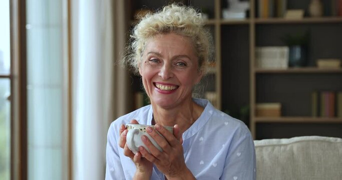 Happy middle aged pretty lady holding cup of hot drink in two both hands, looking at camera with toothy smile, laughing, enjoying mug of morning coffee, tea, sitting on couch at home