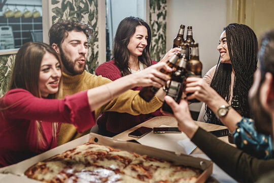 Multi-ethnic happy friends celebrating raising beer bottles having fun dining at pizza party in home interior - food and drink people lifestyle concept