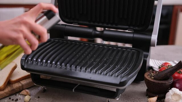 Woman greasing home electric grill surface with vegetable oil using oil spray