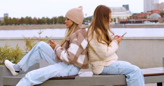 Modern addictions - two young women sit buried in smartphones during an outdoor walking in autumn time.