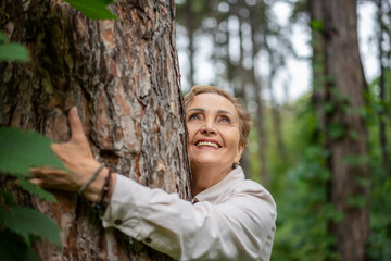 Portrait beautiful happy caucasian active senior woman enjoying nature hugging pine trunk in forest