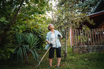 Beautiful happy active senior woman pensioner cleaning fallen leaves using a rake in the yard of a country house in autumn