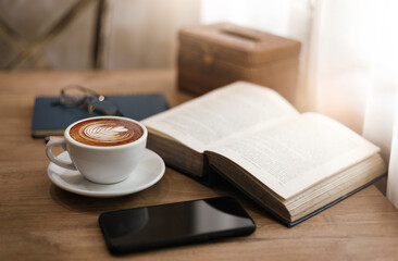 Close up view, Latte coffee in white cup with old book and smart phone on wooden table near bright window. blurred background, eyeglasses on blue note book and pen, vintage color tone