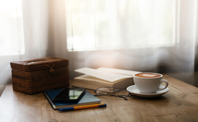 Close up view, Latte coffee in white cup with old book and smart phone on wooden table near bright window. blurred background, eyeglasses on blue note book and pen, vintage color tone