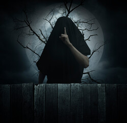 Grim reaper showing silence sign with finger standing over dead tree, full moon and cloudy spooky...