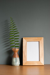 Light wooden frame mockup with fern leaves in a ceramic vase on a wooden table and grey background. blank space, vertical image, still life, copy space.