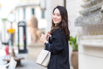 Portrait of a beautiful long-haired Asian woman in a black coat, carrying a walking bag, smiling in a good mood in the city outdoors.