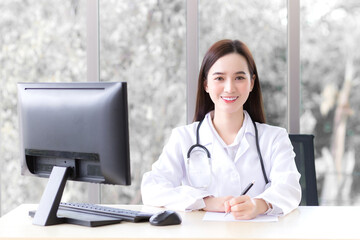 Asian beautiful woman doctor who wears medical coat is working in office room at hospital while has computer put on table.