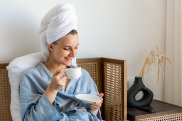 a young woman in a blue bathrobe and a white towel on her head lies on the bed of a hotel room and drinks fragrant coffee. Great start to the day. Morning routine concept - obrazy, fototapety, plakaty
