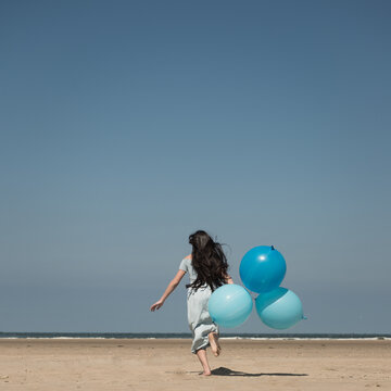 fine art portrait of girl in blue dress running with balloons on beach