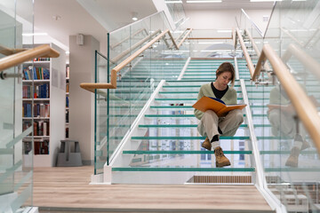 Focused mature female reader sits on modern glass stairs step enjoying interesting book in contemporary public library. Curious woman in casual outfit reads about world history getting new knowledge