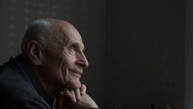 Wrinkled face of smiling senior man sitting at table in dark room and looking to window side view