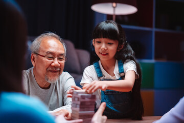 Asian happy family of little daughter play toy wooden block in home. Grandparents and young woman loving couple parent having fun play stack tower wood block game at night in living room.