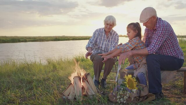 Affectionate old grandparents care little lovely granddaughter warming hands sitting near bonfire on lake shore at sunset outdoors. Funny calm family relax, happy carefree childhood at summer holiday