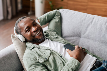 Satisfied middle aged african american man in casual, in wireless headphones enjoys favorite music with phone
