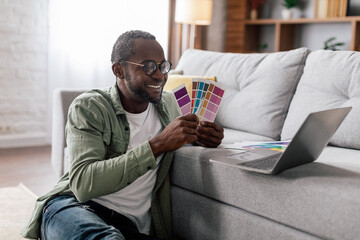 Cheerful adult black businessman designer in glasses and casual show color palettes in laptop in living room