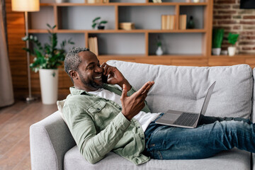 Smiling middle aged african american male in casual calling by phone, work with laptop, sit on sofa