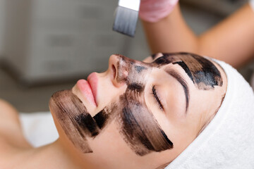 cosmetologist applying black mask on pretty woman face wearing black gloves, gorgeous woman in spa...