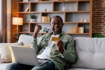Glad excited adult black man in glasses and casual rejoices in victory with computer and credit card
