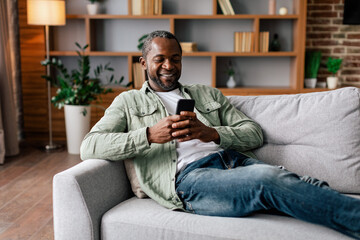 Glad mature african american male in casual resting and relaxing, chatting on smartphone, watching video