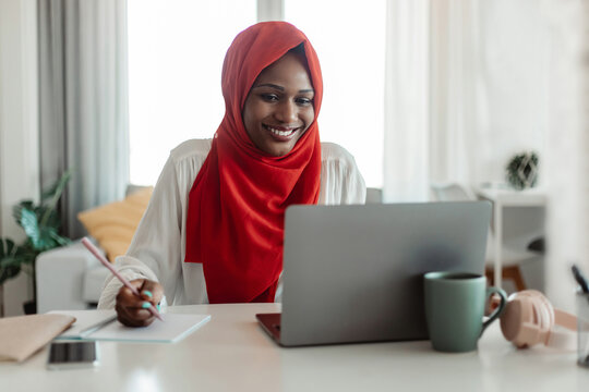 Positive Muslim African American Woman Sitting At Workplace, Using Laptop And Writing In Notebook, Home Office