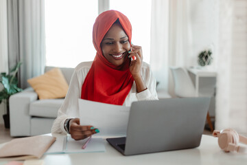 Positive black muslim businesswoman working at home office, holding documents and having phone...