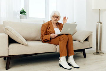 pleasant, stylish elderly lady in white boots and brown suit is sitting in a bright interior, keeping in touch with loved ones at a distance, communicating with them via video link. Full-length photo