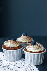 Three delicately delicious muffins with butter cream and blueberries on a lacy napkin on a dark table. Delicious dessert. front view