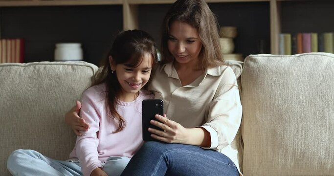 Cheerful happy mom and daughter child talking on video call on smartphone, watching online content, shopping on Internet, smiling, laughing, having fun, sitting on home couch together, hugging