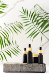 Concrete black podium for cosmetic products close-up among tropical leaves on the background of a marble white wall, a black bottle with a pipette and a jar of cream