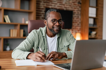 Smiling middle aged african american male in glasses and casual working on laptop in home office interior - Powered by Adobe