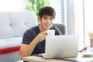 Happy Asian casual man working with laptop holding a cup of coffees, sitting on the floor in the house.