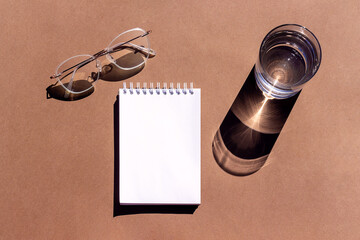 Empty notepad, glass of water and eyeglasses with strong shadows on brown background. Creative mockup. Top view, flat lay, Still life