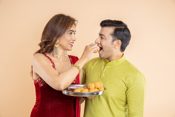 Happy young indian couple celebrate diwali festive, Woman put laddu or laddoo sweet in man mouth and having fun together, isolated studio background.