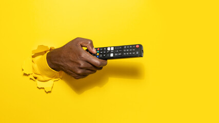Black male hand holding remote control through the torn yellow paper background, pointing it aside...