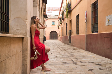 Obraz na płótnie Canvas Young, attractive, blonde woman, wearing an elegant red party dress and holding golden high heels in her hand, barefoot, leaning against the wall of a city alley. Concept beauty, fashion, elegance.