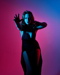 Naklejka premium Close up portrait of beautiful woman model wearing elegant black dress and crown, posing against a studio background with fantasy inspired arm gestures, multi coloured creative lighting.