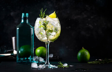 Gin tonic lime alcoholic cocktail drink with dry gin, rosemary, tonic and ice in big wine glass. Black bar counter background, steel bar tools - obrazy, fototapety, plakaty