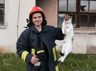 Close-up portrait of heroic fireman in protective suit and red helmet holds saved cat in his arms. Firefighter in fire fighting operation.