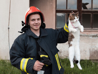 Close-up portrait of heroic fireman in protective suit and red helmet holds saved cat in his arms....