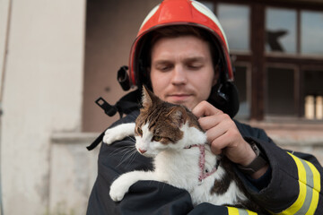 Close-up portrait of heroic fireman in protective suit and red helmet holds saved cat in his arms....