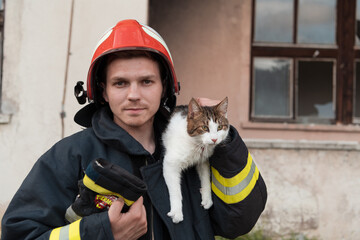 Close-up portrait of heroic fireman in protective suit and red helmet holds saved cat in his arms....