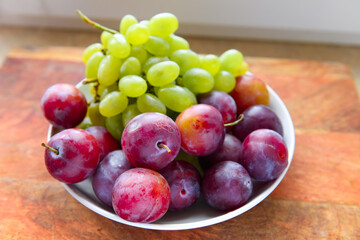 green grapes and plums in a plate on a wooden board, windowsill, concept of fresh fruits and healthy food