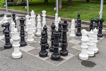 A chessboard on asphalt with white and black chess pieces for playing and competing in a chess battle on the square in front of the City Administration building near the park and a bench for fans.